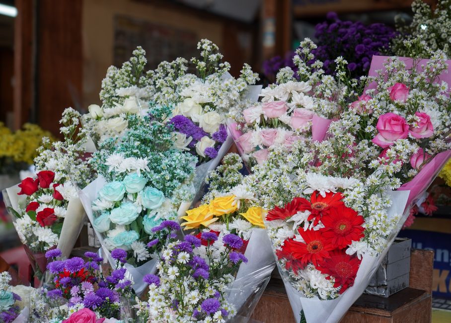 A vibrant display of floral bouquets featuring a variety of fresh flowers arranged in wrapping paper and tissue paper. There are bouquets with pink roses, red gerberas, yellow roses, and blue roses, c
