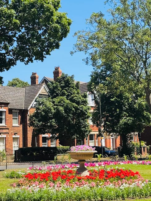 A vibrant flower bed featuring red, pink, and white blooms situated in a landscaped park setting with lush green grass and a decorative stone planter filled with pink and purple flowering plants. Surr
