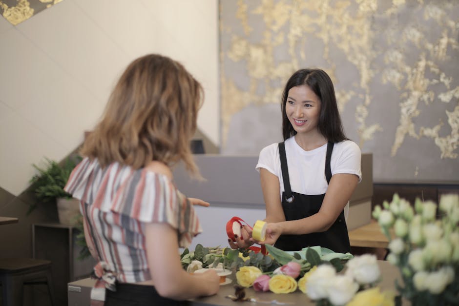 A floral arrangement is being prepared at Florist Harold Hill, with a young female florist wearing a black apron over a white T-shirt, smiling as she hands over a small wrapped bouquet featuring vibra