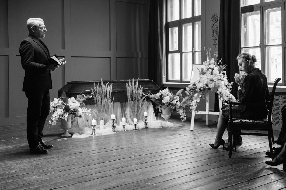 A woman seated in a dark upholstered chair during a funeral ceremony, holding and biting a flower, with a large floral arrangement featuring white roses, lilies, and greenery placed on an easel beside