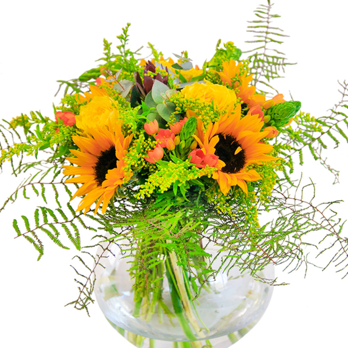 The essence of Harold Hill's sunniest seasons is captured in this lush, striking bouquet, arranged with an expert's eye for colour and balance. Towering above an array of rich foliage, radiant sunflowers stand proud-petals glowing in warm gold and orange hues, centers deep and inviting, like a summer day basking in sunlight at Central Park. Surrounding them, yellow garden roses unfurl in tightly packed whorls, adding luxurious detail and softness. Scarlet hypericum berries are scattered throughout, their vivid poppy-red beads offering both contrast and a sense of playful energy, as if sprinkled by nature herself. Cascading gently over the sides, wispy fern fronds and textured foliage offer movement and echo the wild, untamed greenery found in the gardens around Harold Hill. Subtle hints of bluish-green eucalyptus and pale golden filler, likely local goldenrod, bring airy freshness and draw the eye around the bouquet. All these elements find harmony in a perfectly clear, rounded glass vase, where each vibrant stem is beautifully displayed through pure water. With every glance, you sense the detailed craftsmanship and passion that went into assembling this showpiece-an arrangement equally suited to an anniversary celebrated near St. Neots Road or an elegant table setting for a family gathering. The bouquet's fragrance is soft but present, mingling rose and green notes, creating an inviting atmosphere wherever it's placed. It's a radiant tribute to the best of Harold Hill's flora and the artistry of its florists.
